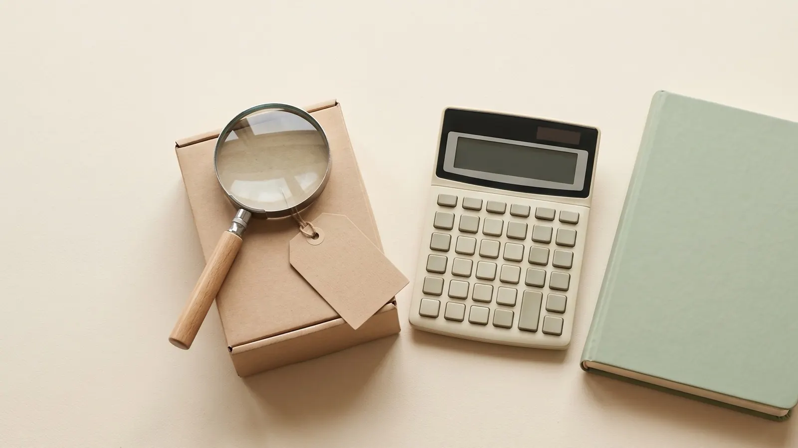 Magnifying glass on a kraft carton with a blank tag beside a calculator and a closed green notebook